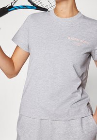 Person iført en lys grå Björn Borg t-shirt og shorts, der holder en sort og blå tennisracket over skulderen.