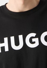 T-shirt en coton noir avec de grandes lettres blanches en caractères gras épelant "HUGO" sur la poitrine. Col rond et texture douce.