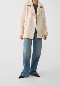 Giacca in shearling beige con colletto ampio, chiusura con zip e due tasche laterali, abbinata a jeans svasati blu sbiaditi e scarpe nere a punta.