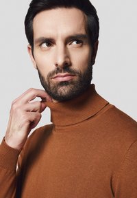 Brązowy sweter z golfem, z prążkowanym kołnierzem i mankietami, miękka faktura, noszony przez mężczyznę dotykającego brody, neutralne tło.