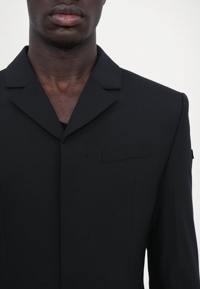 Blazer nera strutturata con colletto a scialle, tasca sul petto singola e senza bottoni. Tessuto elegante con una consistenza liscia, design minimalista.