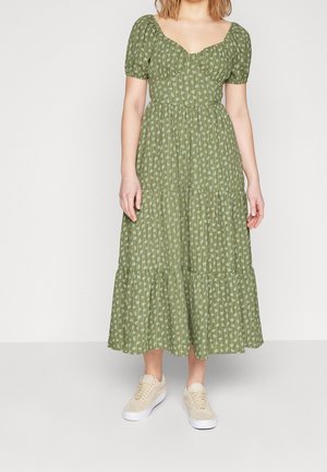 Donna che indossa un vestito midi verde a maniche corte a balze con una piccola stampa floreale e sneakers beige, in piedi davanti a uno sfondo semplice.