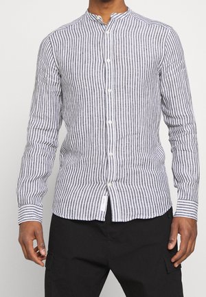 Man draagt een slim-fit, langermig, zwart-wit verticaal gestreept overhemd met knopen en een mandarijnkraag, gecombineerd met een zwarte broek.