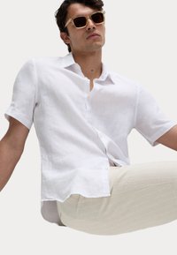 Camisa de lino blanca de manga corta con tapeta de botones, corte relajado y tejido texturizado. Combinada con pantalones a rayas en un tono beige claro. Lleva gafas de sol doradas.