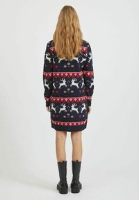 Robe-pull bleu marine avec un motif de renne blanc et des accents rouges. Manches longues, poignets côtelés, et longueur au genou. Bottes plateformes noires.