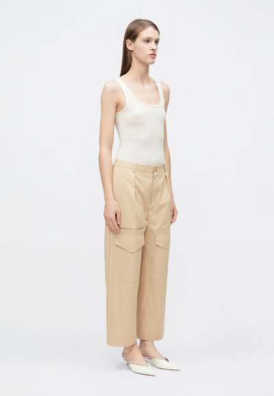 Λεπτή γυναίκα που φοράει μπεζ τοπ χωρίς μανίκια, φαρδιά μπεζ παντελόνια cargo και λευκά παπούτσια mule με μυτερή μύτη, στέκεται μπροστά σε απλό λευκό φόντο.