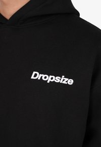 Zwarte hoodie met witte "Dropsize" tekst gedrukt op de linkerkant van de borst, toont bovenlichaam en gedeeltelijke hals.