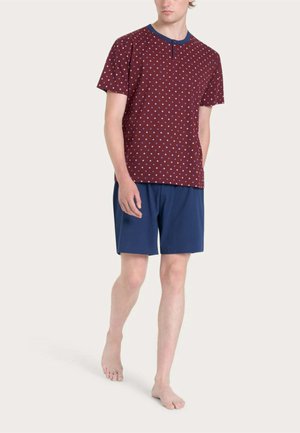 Giovane uomo che cammina a piedi nudi indossando una camicia bordeaux a maniche corte con piccoli puntini blu e bianchi e pantaloncini blu navy su uno sfondo uniforme.