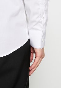 Biała koszula zapinana na guziki o gładkiej fakturze; posiada zaokrąglony dół i mankiet zapinany na pojedynczy guzik. W tle widoczne czarne spodnie.