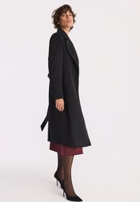 Kobieta ubrana w długi czarny płaszcz na bordowej sukience, czarnych półprzezroczystych rajstopach i czarnych szpilkach, stojąca na tle białym.