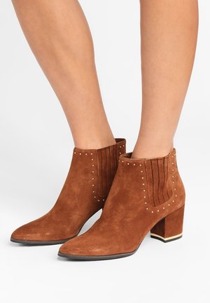 Bottines en daim marron avec talons blocs, panneaux latéraux élastiques et petits clous dorés portées sur des jambes nues.