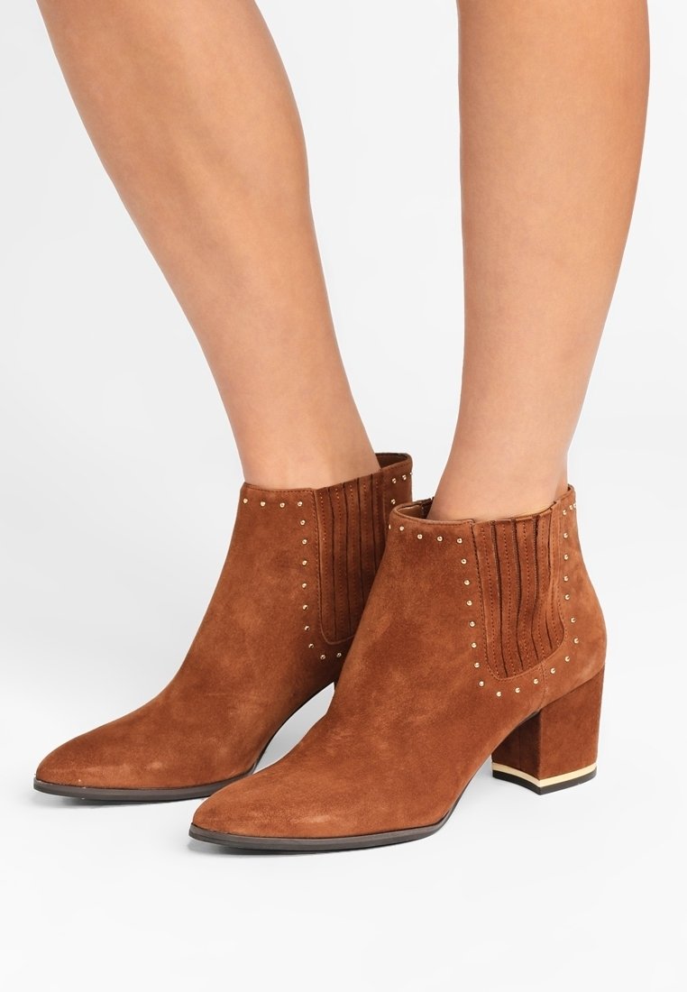 Bottines en daim marron avec talons blocs, panneaux latéraux élastiques et petits clous dorés portées sur des jambes nues.