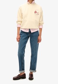Felpa corta color crema con ricami rosa, indossata su una maglietta rosa chiaro, abbinata a jeans dritti blu e mocassini bordeaux.