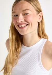 Hvid ribbet tanktop med høj hals og armhuller. Stoffet ser blødt ud med en let tekstur. Håret er langt og glat.