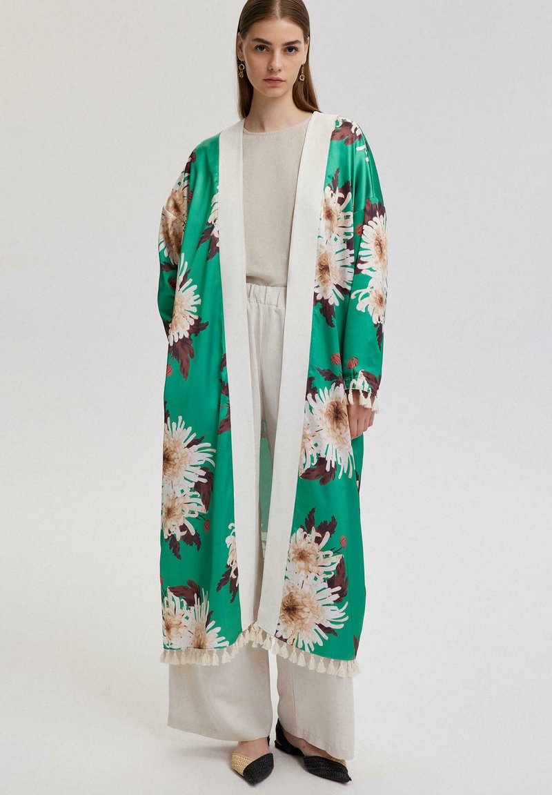 Roheline lilleline kimono, millel on suured valged ja pruunid lillemustrid, mille ääres on kindel valge serv. Ühendatud heledate pükste ja terava ninaga kortsukatega.