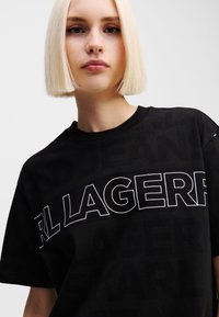 Crna pamučna majica s velikim bijelim natpisom "KARL LAGERFELD" i suptilnim uzorkom u pozadini. Opušteni kroj, klasična okrugla izreza.