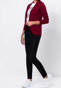 Burgunderfarbener Cardigan mit strukturierter Oberfläche und fallendem Vorderteil, kombiniert mit engen schwarzen Hosen und weißen Sneakern. Das Outfit wird von einem schlichten Graphic-T-Shirt ergänzt.