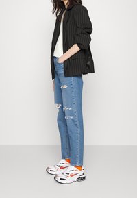 Blazer negro a rayas sobre una blusa blanca, combinado con jeans azules desgastados y zapatillas blancas con acentos naranjas y suelas negras.