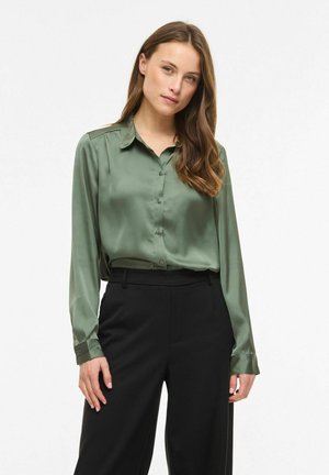 Femme aux longs cheveux bruns portant un chemisier en satin vert boutonné, rentré dans un pantalon noir taille haute, debout devant un fond blanc.