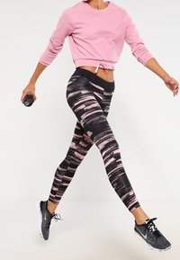 Osoba w czarnych i różowych legginsach w wzory, różowej kroplonej bluzie oraz czarnych butach sportowych, w biegu trzymająca hantle w lewej ręce.