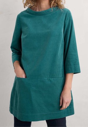 Femme portant une tunique bleu sarcelle à manches longues avec une poche avant, main partiellement dans la poche, sur un fond clair uni.
