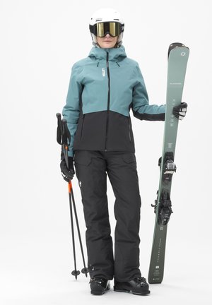 Tenue de ski avec une veste turquoise et noire, un pantalon noir, des bâtons de ski et des skis. Les skis sont verts avec une surface texturée et des fixations.