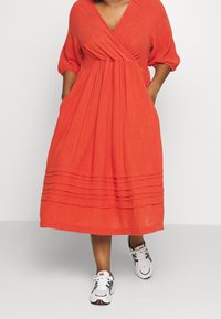 Robe en lin rouge avec un design à enroulement, manches courtes bouffantes et plis horizontaux au bas, accompagnée de baskets de sport blanches et grises.
