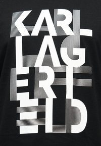 Λευκό έντονο κείμενο "KARL LAGERFELD" με κεφαλαία γράμματα σε μαύρο ύφασμα, με οριζόντιες ριγέ γραμμές ενσωματωμένες στα γράμματα.