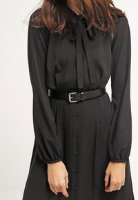 Robe noire à manches longues avec un col noué et une fermeture boutonnée sur le devant, cintrée par une large ceinture noire avec une boucle argentée. Texture de tissu lisse.