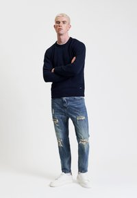 Granatowy sweter z dzianiny, o lekkiej fakturze, z okrągłym dekoltem. Noszony z przetartymi niebieskimi dżinsami, wykończony białymi trampkami.