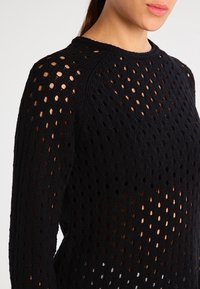 Svart stickad tröja med ett perforerat mönster med cirkulära hål, rundad halsringning och långa ärmar, som visar en strukturerad tyg.