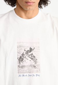 Hvid bomulds t-shirt med et sort-hvidt grafisk motiv af to hunde, der leger, med teksten "Al arbejde og ingen leg" i blå under billedet.