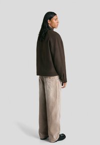 Rjava oversize jakna z gumbnim ovratnikom, kombinirana z bež korduroj hlačami. Model stoji obrnjen stran, obut v črne čevlje.