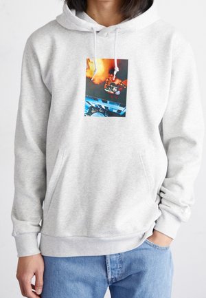 Person trägt einen hellgrauen Hoodie mit einer farbigen Grafik eines oberkörperfreien Mannes, der eine Torte und eine brennende Fackel über einem blauen Gebäude hält.