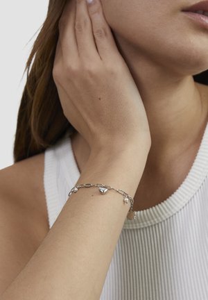 Silver chain bracelet with heart charm and small bead accents, worn on a wrist. Background features a textured white top.