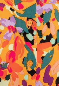 Bunt gemusterter Stoff in Orange mit lila, grünen und schwarzen Motiven. Zwei orangefarbene Knöpfe an der Vorderseite setzen Akzente. Strukturierter Baumwollstoff.