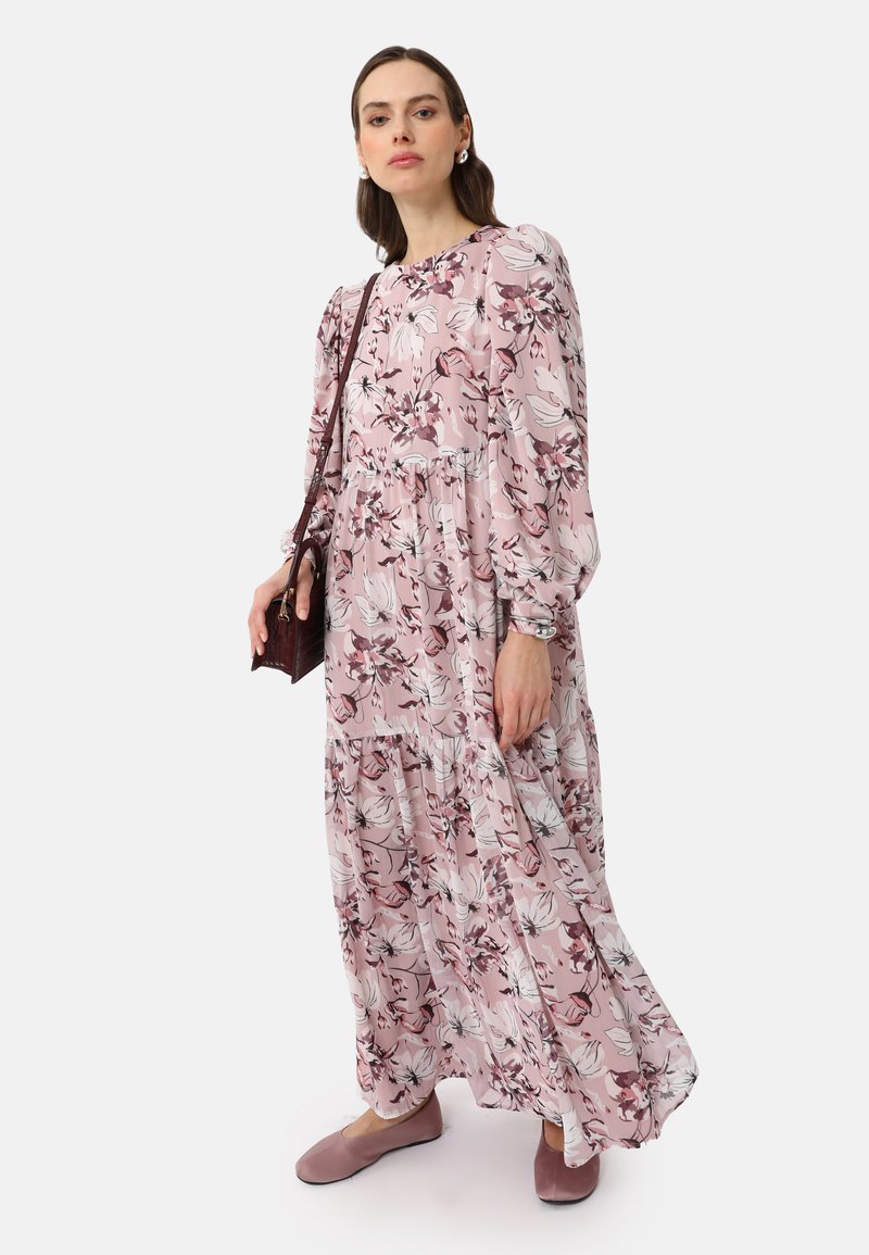 Robe longue rose à motifs floraux avec des manches bouffantes, un design en étages et des délicats motifs de fleurs blanches. Associée à des chaussures roses douces et un petit sac.