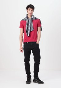 Rotes Kurzarmhemd, schwarze Jeans, schwarze Sneakers. Gestreifter schwarz-weißer Pullover lässig über den Schultern drapiert. Model steht vor einem schlichten Hintergrund.
