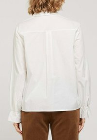 Blouse blanche à manches longues avec un plissé dans le dos. Présente des accents en dentelle sur les poignets et est confectionnée dans un tissu lisse et léger.
