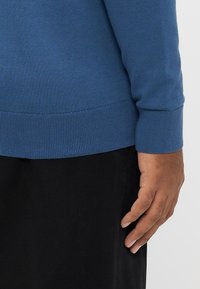 Niebieski sweter z prążkowanymi mankietami i ściągaczem u dołu, wykonany z gładkiej dzianiny. U dołu widoczne czarne spodnie, podkreślające kontrast kolorów.