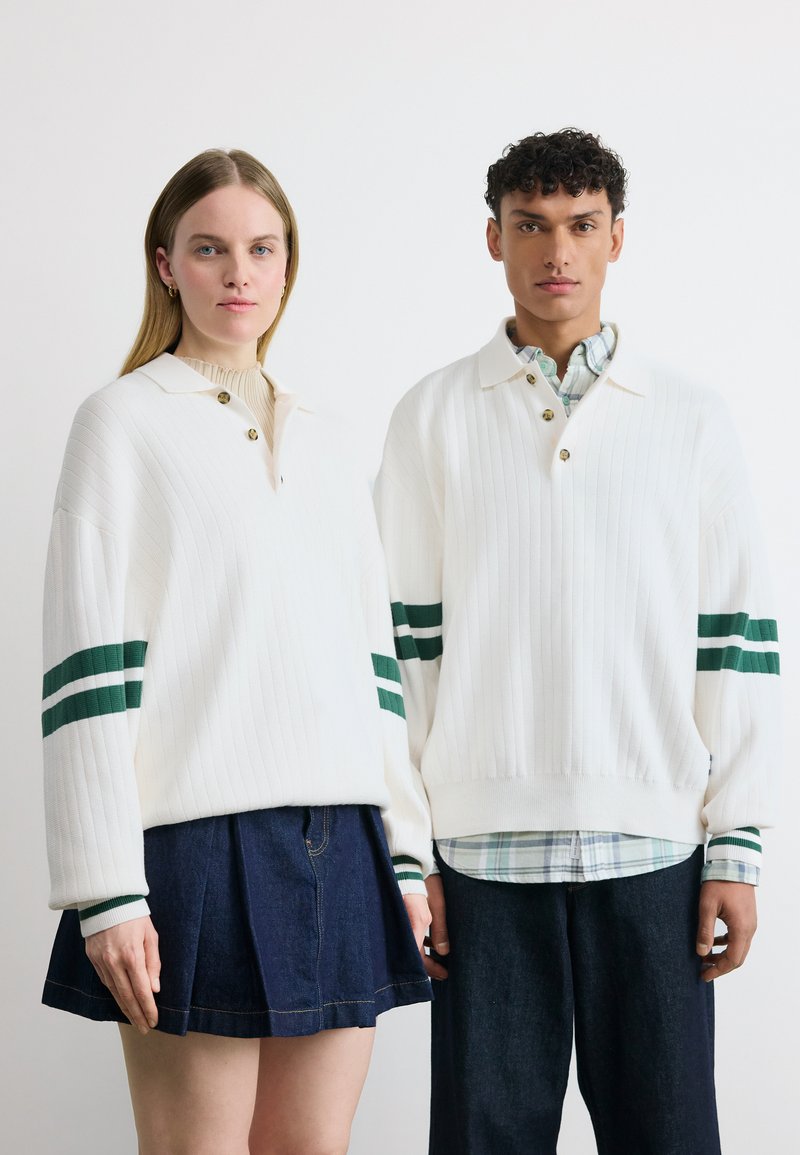 Δύο μοντέλα φορούν λευκά ριμπ πλεκτά πουλόβερ με πράσινες ρίγες στα μανίκια· η γυναίκα το συνδυάζει με τζιν φούστα, ο άνδρας με σκούρο παντελόνι.