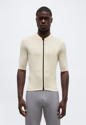 Mann iført beige sykkeltrøye med korte ermer og frontglidelås, samt grå polstrede sykkelshorts, stående foran en ensfarget hvit bakgrunn.