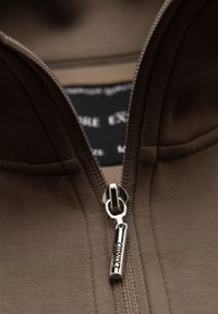 Chaqueta marrón con cremallera, que presenta una textura suave y un tirador de cremallera plateado, con una etiqueta de marca visible en el área del cuello.