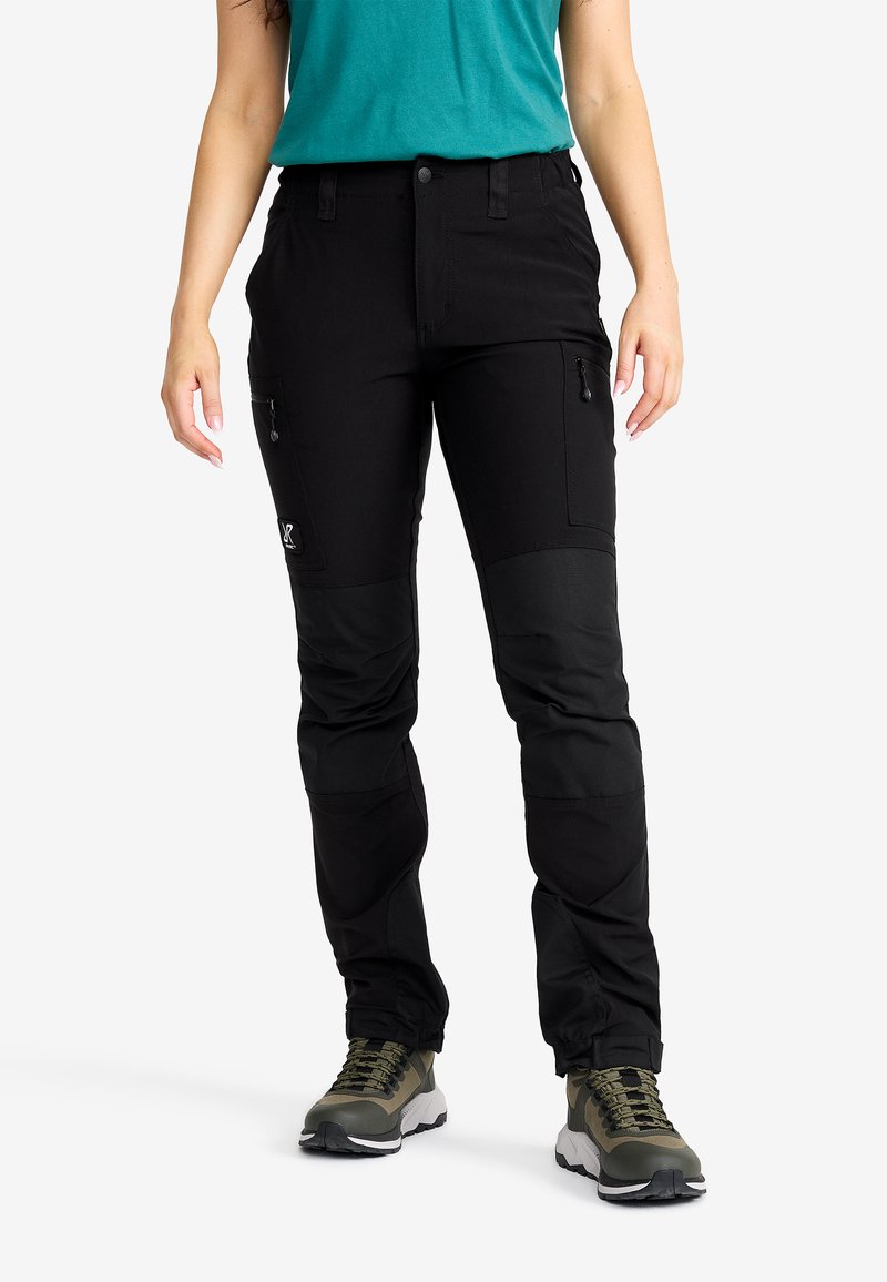 Person som bär svarta cargobyxor med dragkedjefickor och olivgröna vandringsskor, står mot en enfärgad vit bakgrund.