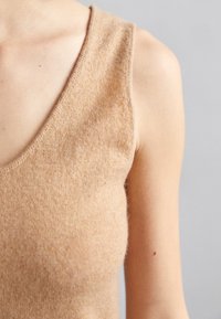 T-shirt sans manches en tricot beige avec un décolleté en V prononcé, présentant une surface douce et légèrement texturée et une coupe ajustée.