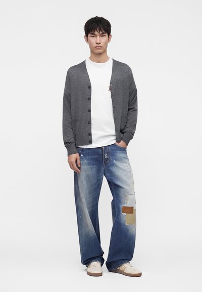 Jeune homme debout, la main dans la poche, portant un cardigan gris boutonné, un t-shirt blanc, un jean large bleu avec des patchs et des baskets blanches.
