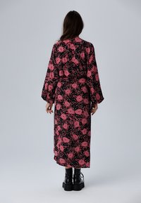 Lång svart klänning med livfullt rosa blommönster, långa ärmar och en åtsittande midja, kombinerat med klumpiga svarta stövlar. Slät tygtextur.