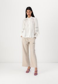 Weiße Bluse mit Spitze und plissierten Ärmeln, kombiniert mit locker sitzenden beige Hosen und lila Riemchenschuhen. Glattes Material, elegantes Design.