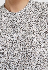 Tissu blanc avec un motif tacheté en brun et noir. Présente un col rond et un détail de bouton au niveau du col. Texture douce.