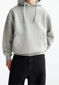 Grijze hoodie van zachte stof met een voorzak, capuchon met trekkoord en geribbelde boorden, gecombineerd met zwarte jeans. Eenvoudig, casual ontwerp.